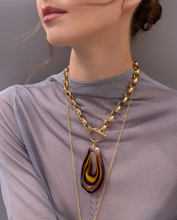A model wearing Katerina Psoma Plume necklace made of a teardrop handmade Murano glass pendant in brown, amber and red colors set on a gold plated thick metal chain with a toggle clasp. 