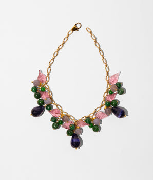 A gold-tone chain necklace with pink and green floral embellishments, featuring faceted green beads, pink enamel leaves, and teardrop-shaped purple and green stones, displayed on a clean white background.